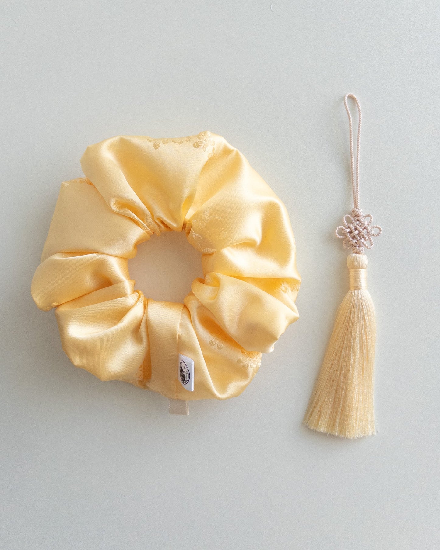 Yellow satin hanbok scrunchie with a decorative Norigae(tassel) on a light gray background