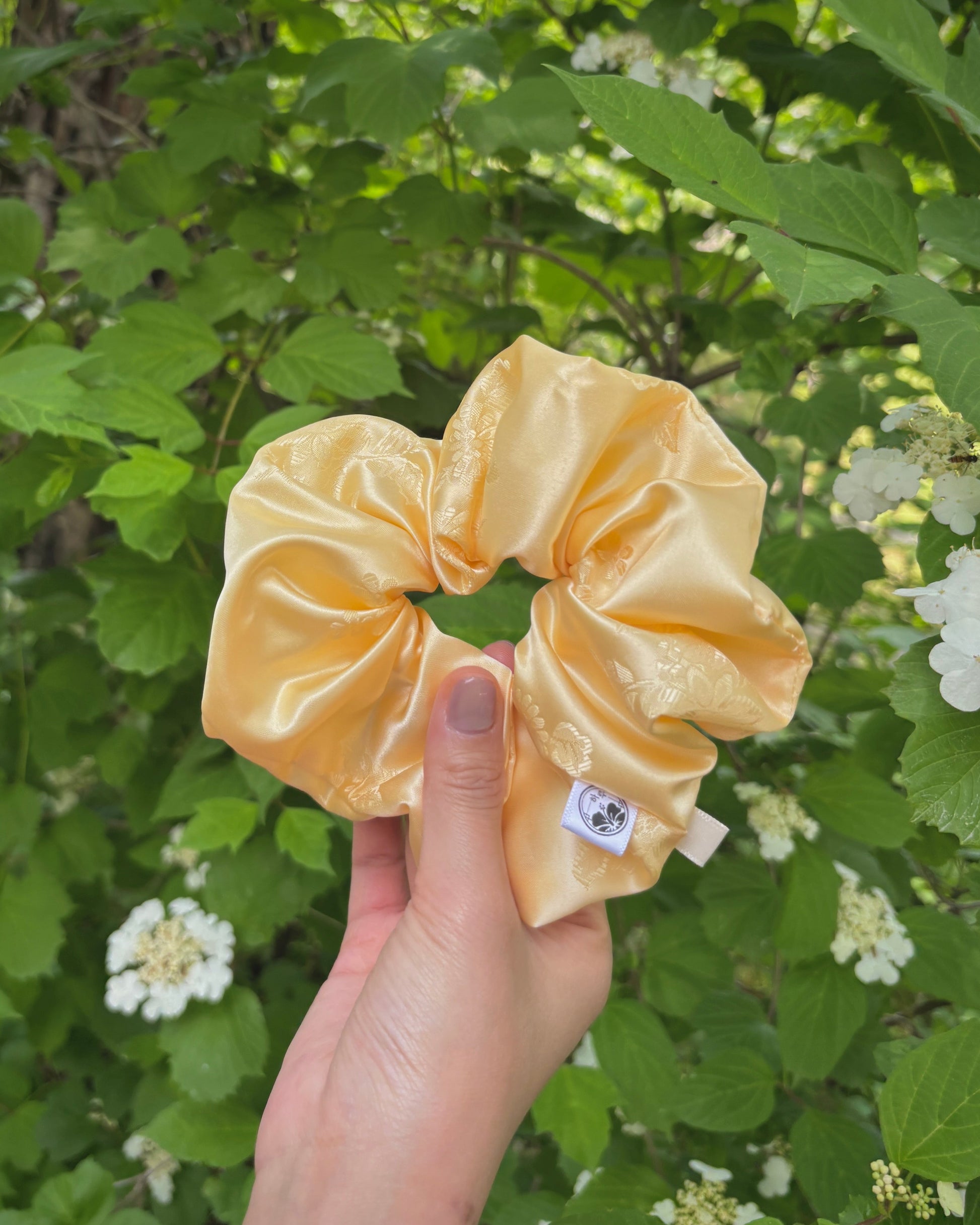 Hand holding a peach-colored scrunchie with a brand label against a green leafy background